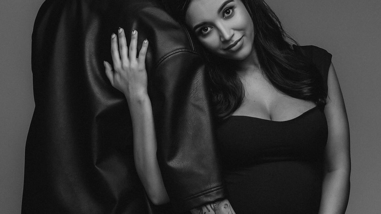 A couple embracing tenderly in a black and white photo. The woman has long dark hair and cradles her pregnant belly. The man has braided hair and wears a leather jacket.