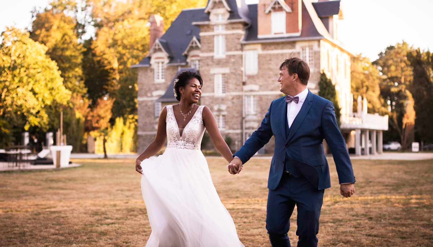 Happy couple walking hand in hand in front of a historic building surrounded by trees. The bride wears a white dress and the groom is in a blue suit during sunset.