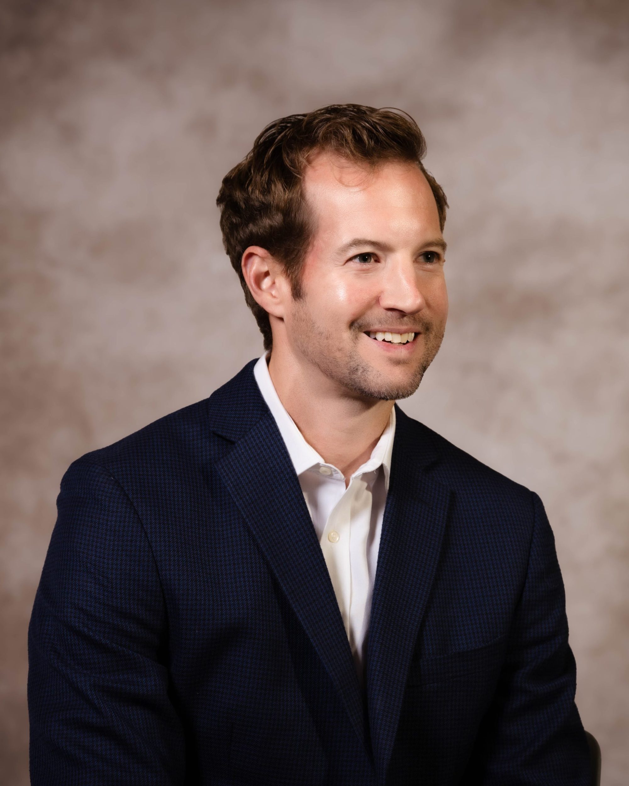 Smiling man in a navy blue suit with a brown textured background looking off-camera. The image conveys a professional and approachable demeanor.