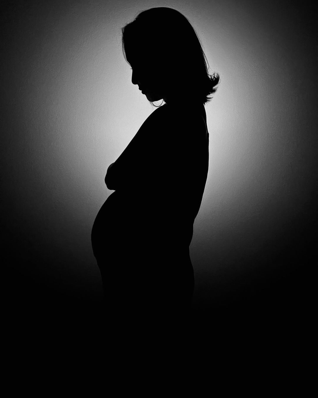 Silhouette of a pregnant woman standing in profile against a softly lit background capturing the beauty and serenity of pregnancy with emphasis on her growing belly.