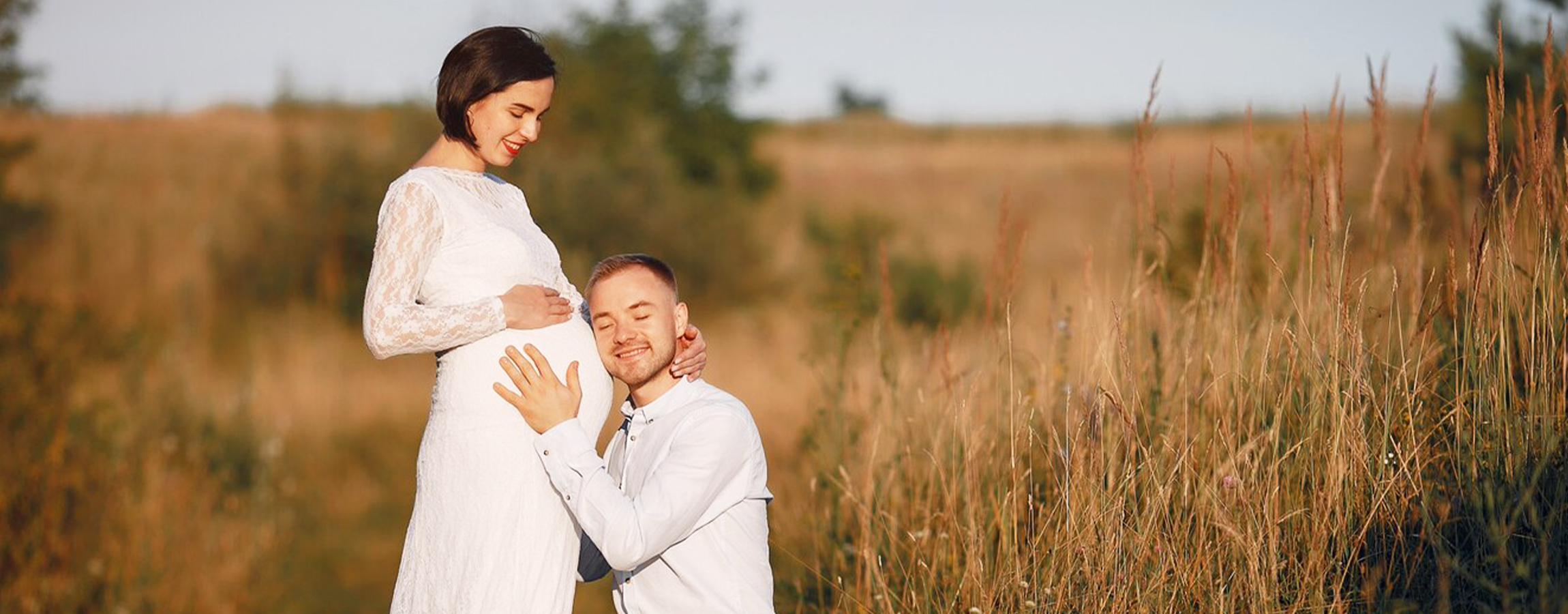 A couple in a field sharing a tender moment as the man kneels and rests his head on the pregnant woman's belly both smiling and dressed in white enjoying the outdoors.