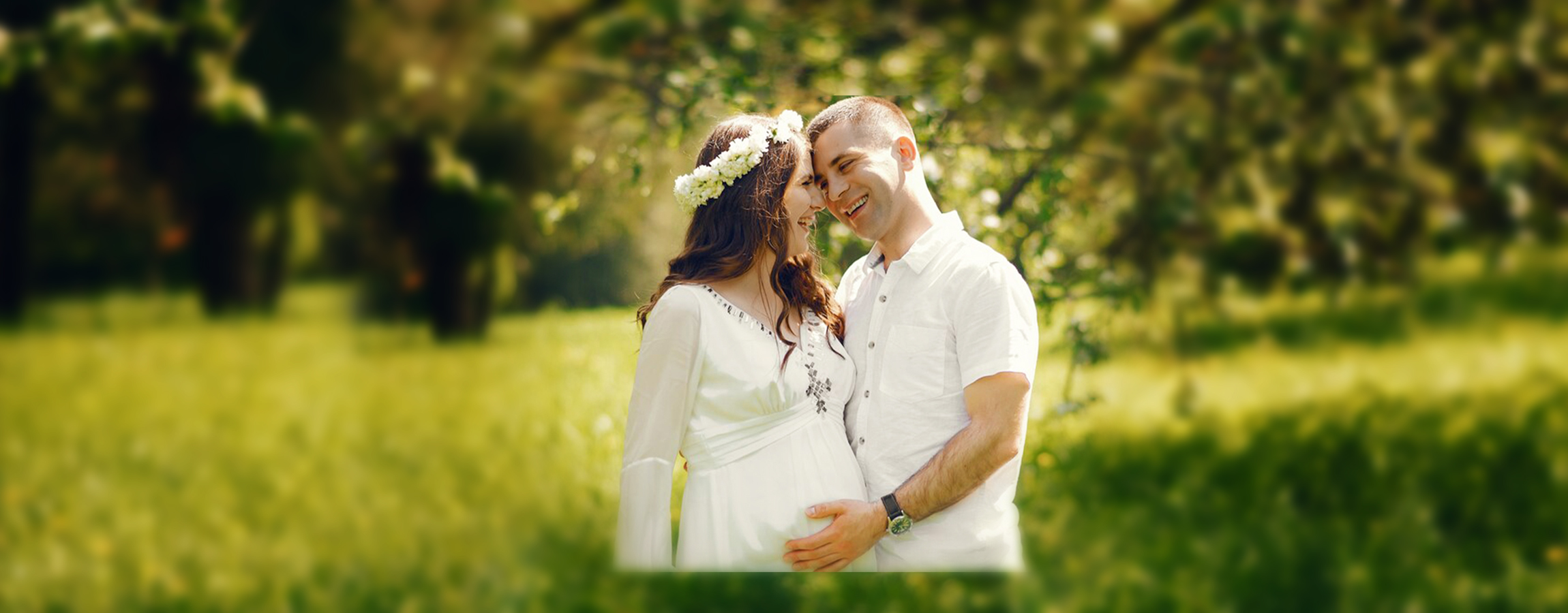 A happy couple in a lush green park shares a moment of affection. The woman is wearing a floral headband and a white dress, while the man lovingly holds her belly.