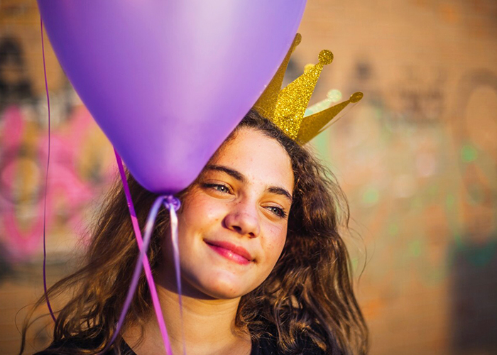 Young girl smiling while holding a purple balloon wearing a golden crown against a colorful graffiti wall. Vibrant and joyful atmosphere on a sunny day.