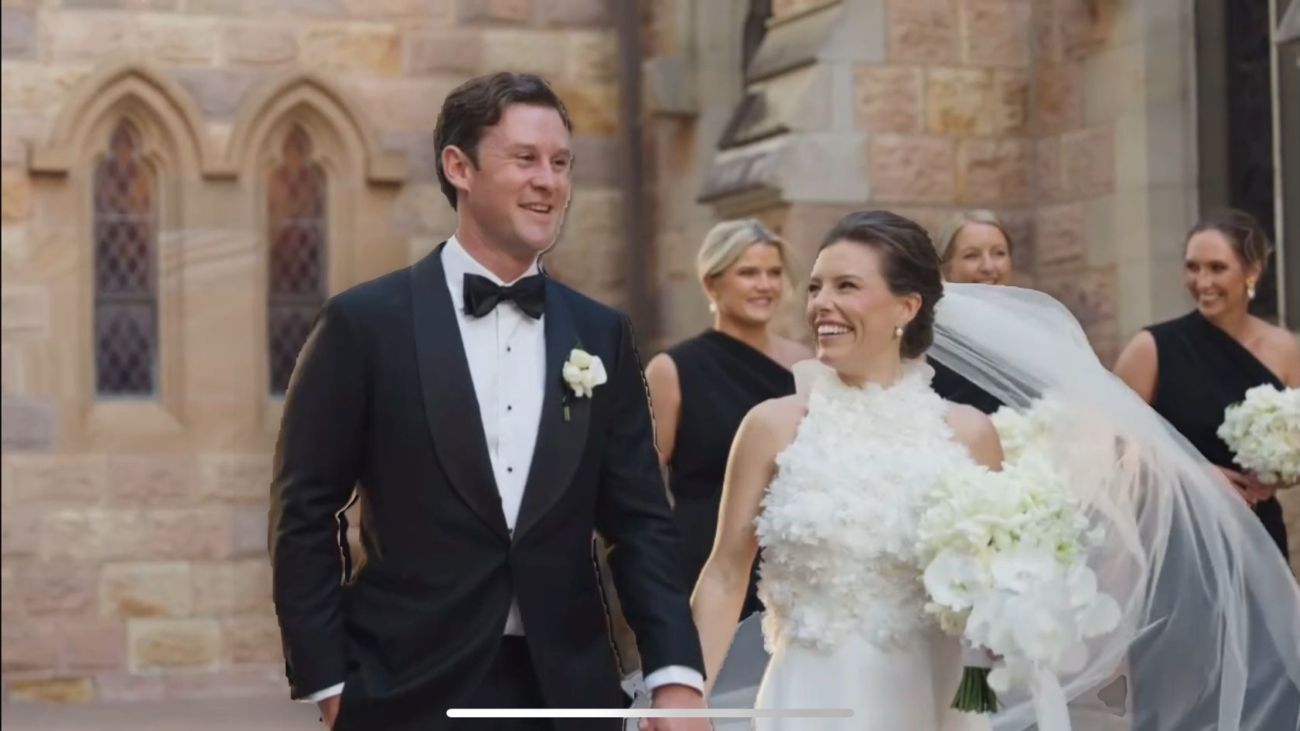 A joyful bride and groom walk hand in hand outside a stone building wearing elegant wedding attire. Bridesmaids in black dresses follow. The bride holds a bouquet of white flowers.