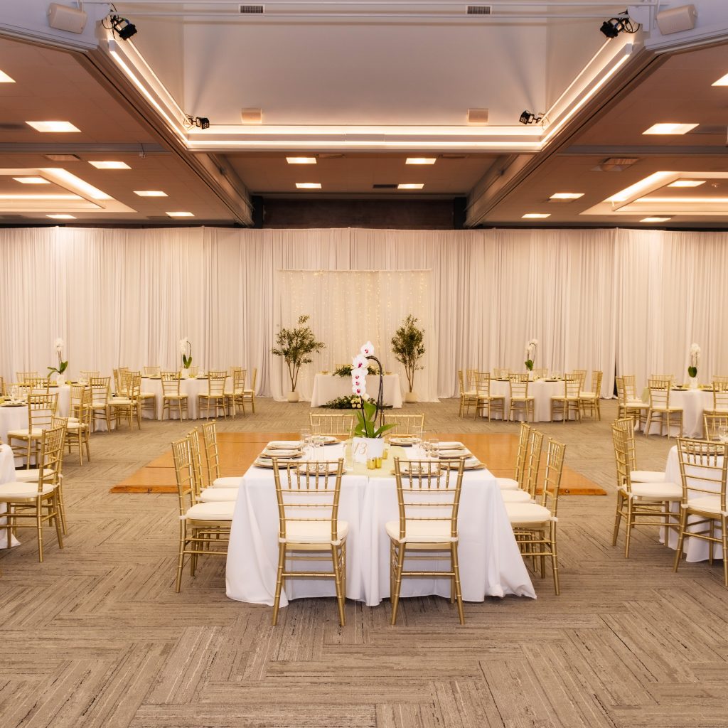 Elegant event hall with round tables decorated in white tablecloths and gold chairs ideal for weddings and corporate events with soft overhead lighting and draped walls.