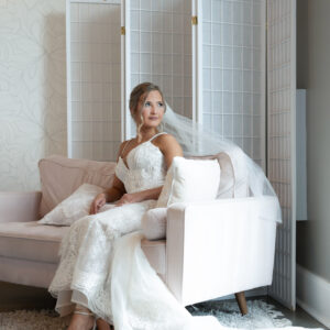 Bride in elegant white wedding dress with lace detail sits on a plush sofa in a softly lit room with a decorative screen creating a serene and romantic setting