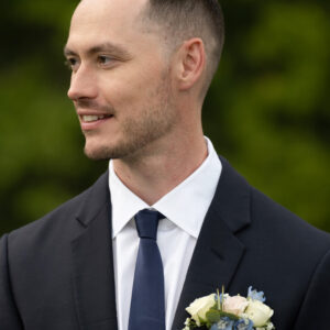 Man in formal suit with flower boutonniere smiles while standing outside against natural green background. Elegant attire and outdoor setting create a sophisticated look.