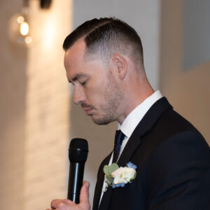Man in a suit with a neat haircut holding a microphone and wearing a boutonniere is standing solemnly against a softly lit background during an indoor event.