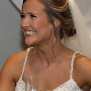 Bride radiantly smiling at a wedding reception wearing a white lace gown and veil. She is seated at a table decorated with elegant glassware under soft ambient lighting.