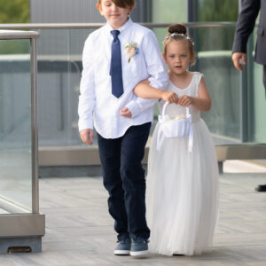 A young boy in a white shirt and tie walks hand in hand with a girl in a white dress holding a small basket, both appear to be part of a wedding ceremony.