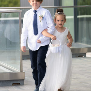 A young boy in a white shirt and tie escorts a little girl in a white dress holding a basket of flowers walking on a terrace with glass railings on a sunny day.