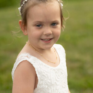 Young girl with brown hair in a bun wearing a white lace dress and floral headband smiles outdoors on a grassy field on a sunny day. Ideal for weddings or celebrations.