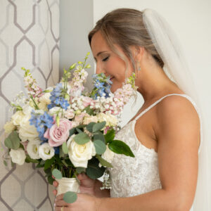 Bride in white lace gown holding pastel floral bouquet smiling near window curtain on wedding day capturing joyful and intimate moment in natural light
