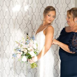 Bride getting ready for her wedding as a woman in an elegant dress helps her. She holds a beautiful bouquet of pastel flowers, standing against a patterned wall.