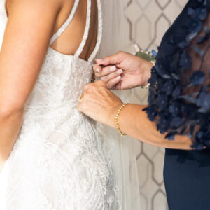 A woman assists in fastening the back of a bridal gown on a bride holding a bouquet of flowers showcasing a special moment during wedding preparations against a patterned background.