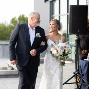 A bride in a white gown walks arm-in-arm with an older man in a suit during an outdoor wedding ceremony. She holds a bouquet of blue and white flowers.
