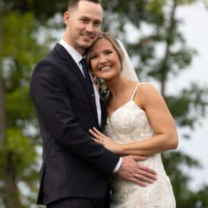 Bride and groom embracing outdoors in a lush green setting on their wedding day capturing a moment filled with love and joy as they smile at the camera.