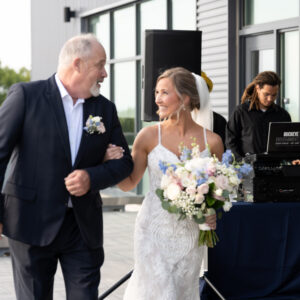 Bride in elegant white gown walks arm-in-arm with a man in a suit holding a bouquet of flowers with a DJ playing music outdoors on a terrace setting with glass doors