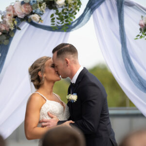 Bride and groom share a romantic kiss under a beautifully decorated floral wedding arch with delicate drapery on a sunny day capturing a special moment of love and joy.