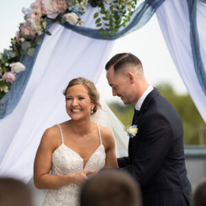Bride and groom exchanging vows under a floral wedding arch on a bright day. The bride is smiling in a white gown while the groom, in a dark suit, holds her hand.