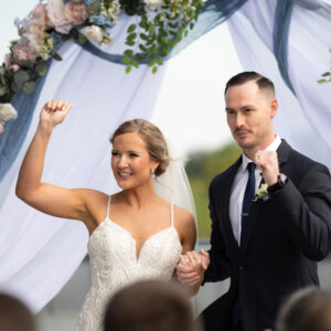 Bride and groom celebrate under a beautifully adorned wedding arch outdoors. The bride wears a white gown and veil while the groom is in a dark suit with a boutonniere.