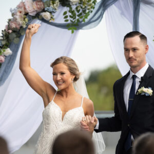 Bride and groom celebrating their wedding under a floral archway with blue and pink accents on a sunny day surrounded by nature and joyous guests.
