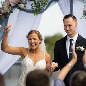 Bride and groom celebrating joyfully under a floral arch at an outdoor wedding ceremony with guests applauding. Bride is raising her hand with a joyful expression.