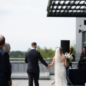 A newlywed couple walks hand in hand at an outdoor wedding reception surrounded by guests enjoying the sunny day and scenic views from the terrace.