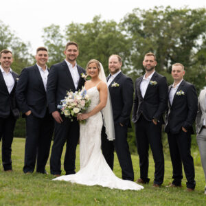 Bride and groom posing outdoors with groomsmen on a grassy field. The bride holds a bouquet while everyone in formal attire smiles against a backdrop of green trees.