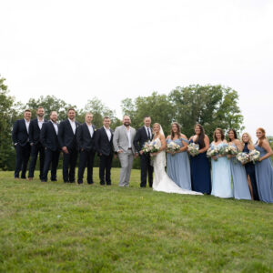 Wedding party posing outdoors in elegant attire on a lush green lawn with trees in the background bride in white gown bridesmaids in blue holding bouquets groomsmen in suits.