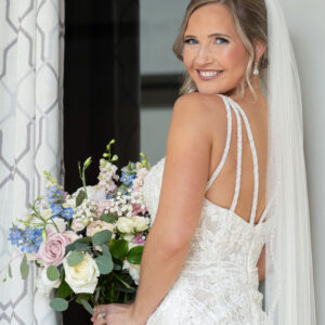 Bride in elegant lace wedding dress holding a bouquet of roses and lilies standing near a window looking over her shoulder with a joyful smile on her wedding day