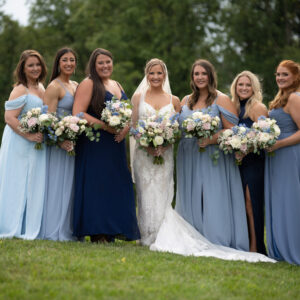 Bride and bridesmaids in elegant blue and white dresses holding floral bouquets pose together outdoors against a lush green background on a wedding day.
