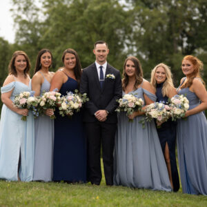 A groom in a dark suit stands with six bridesmaids in matching blue dresses, holding bouquets, against a lush green outdoor backdrop on a wedding day.