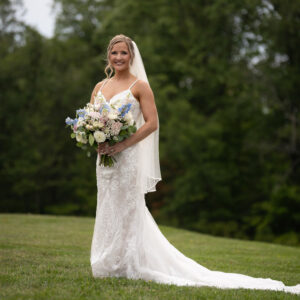 Smiling bride in a white lace wedding dress holding a bouquet of flowers stands on a lush green lawn with a backdrop of tall trees and natural scenery.