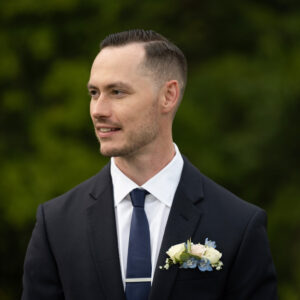 A man in a formal dark suit with a white shirt and navy tie stands outdoors with a green blurred background. He is wearing a boutonniere featuring white flowers.