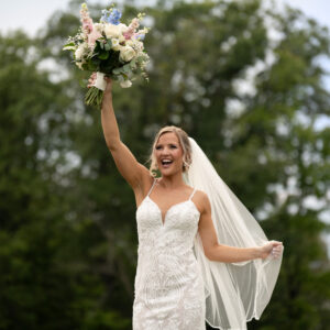 Bride in elegant white gown and veil joyfully lifts floral bouquet against a lush green outdoor backdrop capturing a timeless wedding moment filled with happiness.