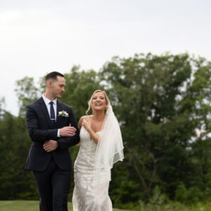 Bride and groom joyfully walk arm in arm on a grassy field surrounded by lush greenery on their wedding day capturing a moment of happiness and celebration outdoors.
