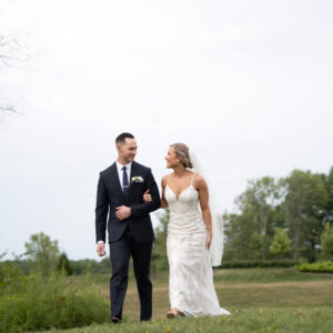 Bride and groom walk together on a grassy field under a cloudy sky capturing a joyful moment in an elegant outdoor wedding setting with lush greenery in the background.