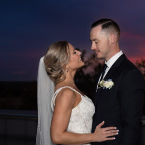 Bride and groom embracing on a rooftop at sunset capturing a romantic moment with the colorful sky in the background highlighting their wedding attire and happy expressions.