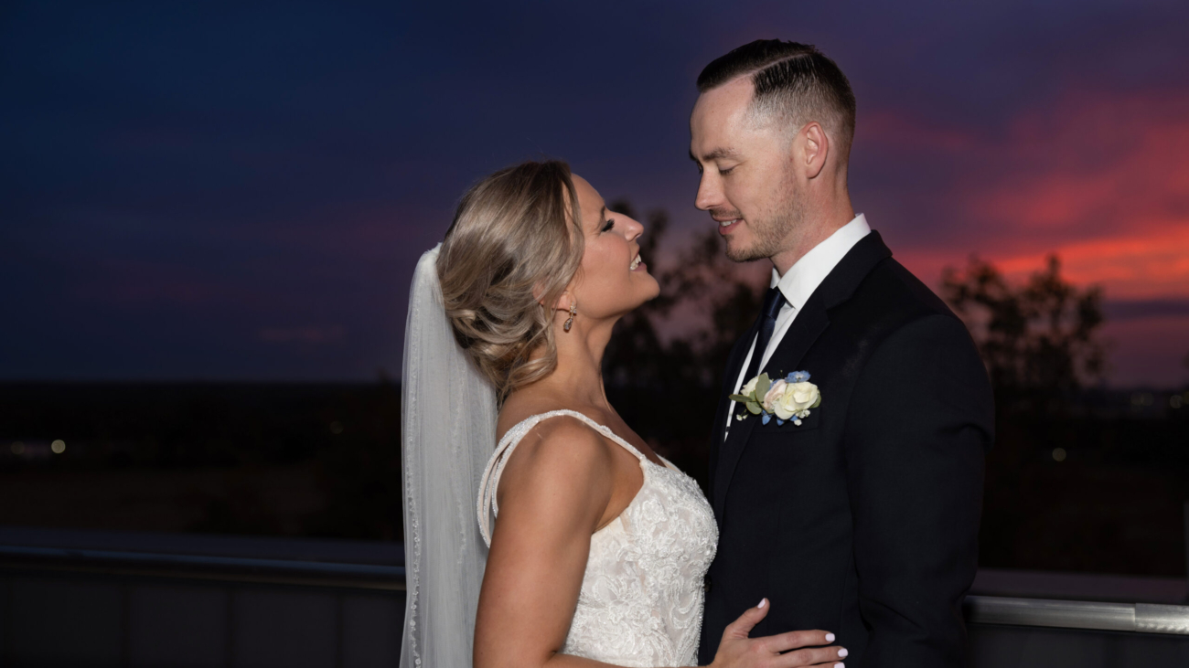 Bride and groom embracing on a rooftop at sunset capturing a romantic moment with the colorful sky in the background highlighting their wedding attire and happy expressions.