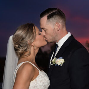 Bride and groom share a romantic kiss at sunset on their wedding day wearing elegant attire with a beautiful pink and purple sky in the background.