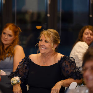 Smiling woman in a blue dress seated at a formal event with other guests dressed elegantly in the background enjoying the occasion in a warmly lit setting