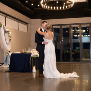 Bride and groom share a romantic first dance in an elegant wedding venue with modern decor, string lights, and a beautifully decorated sweetheart table in the background.