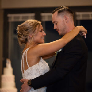 A bride in a white dress and a groom in a black suit share a romantic dance at their wedding reception with a tiered cake in the background against a softly lit setting.