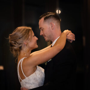 Bride and groom share a romantic first dance at their wedding reception with a tiered cake visible in the background. The couple gazes at each other lovingly.