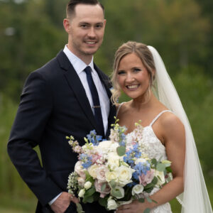 Bride and groom smile joyfully during outdoor wedding ceremony with lush greenery backdrop. Bride holds bouquet of pink white and blue flowers. Groom wears elegant suit.