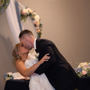 Bride and groom share a romantic kiss at their wedding ceremony with elegant floral decorations and draped fabric creating a beautiful and intimate backdrop.