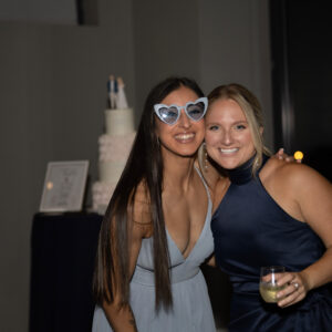 Two women smiling at a party one wearing cat-eye sunglasses and a light blue dress the other in a dark blue dress holding a drink with a decorated cake in the background.