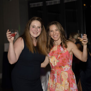 Two women enjoying a night out at a social event each holding a drink and smiling at the camera one wearing a navy dress and the other wearing a floral dress.