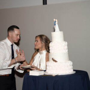 Elegant wedding celebration with a couple standing by a tiered white cake adorned with a small topper on a blue tablecloth creating a joyful and memorable moment.